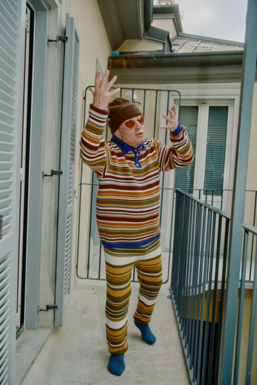 Elderly man in striped outfit raises arms on a balcony with grey railings and a white building facade with shuttered windows in the background.