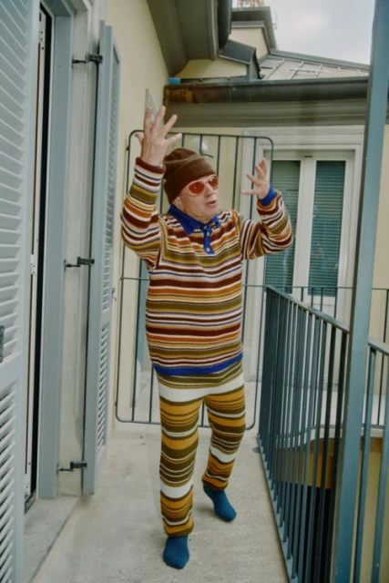 Elderly man in striped outfit raises arms on a balcony with grey railings and a white building facade with shuttered windows in the background.