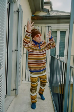 Elderly man in striped outfit raises arms on a balcony with grey railings and a white building facade with shuttered windows in the background.