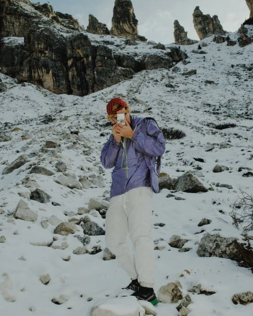Man in jacket taking a photo on a snow-covered mountain with rock formations. The sky is visible in the background.