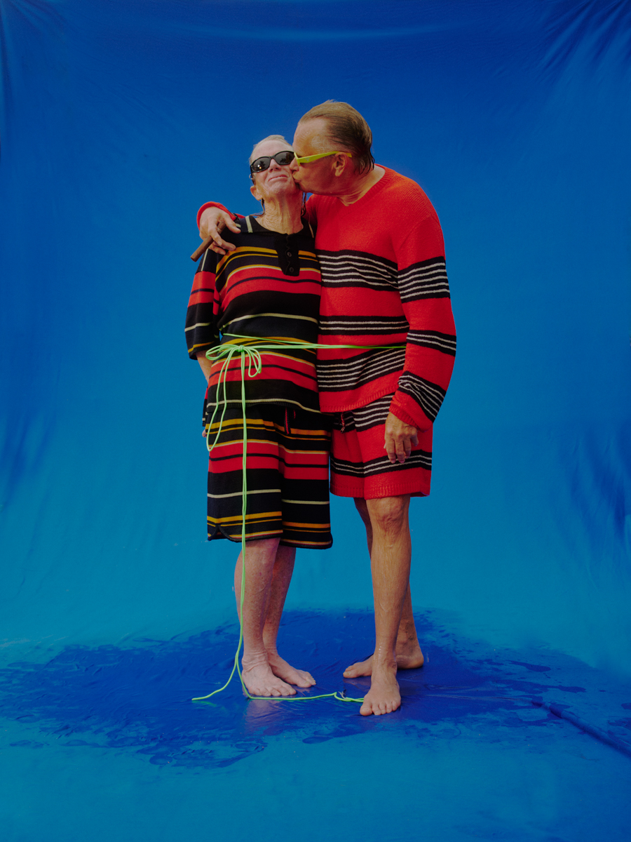 Mature couple in matching striped outfits posing in a studio with a blue backdrop and reflective floor, one kissing the other's cheek.