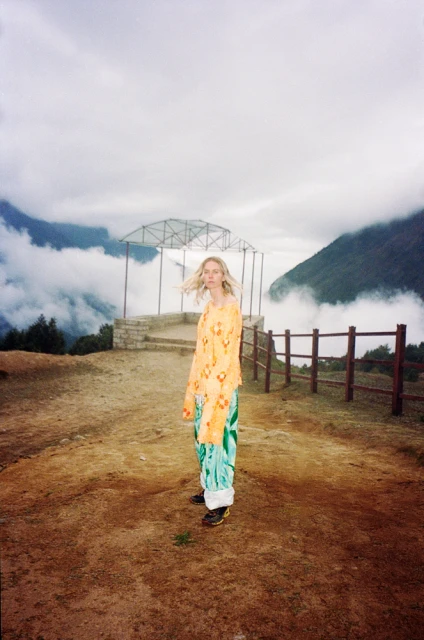 Woman stands on a mountaintop overlook with a metal pavilion and misty mountains in the background, wearing an orange floral top and patterned pants.