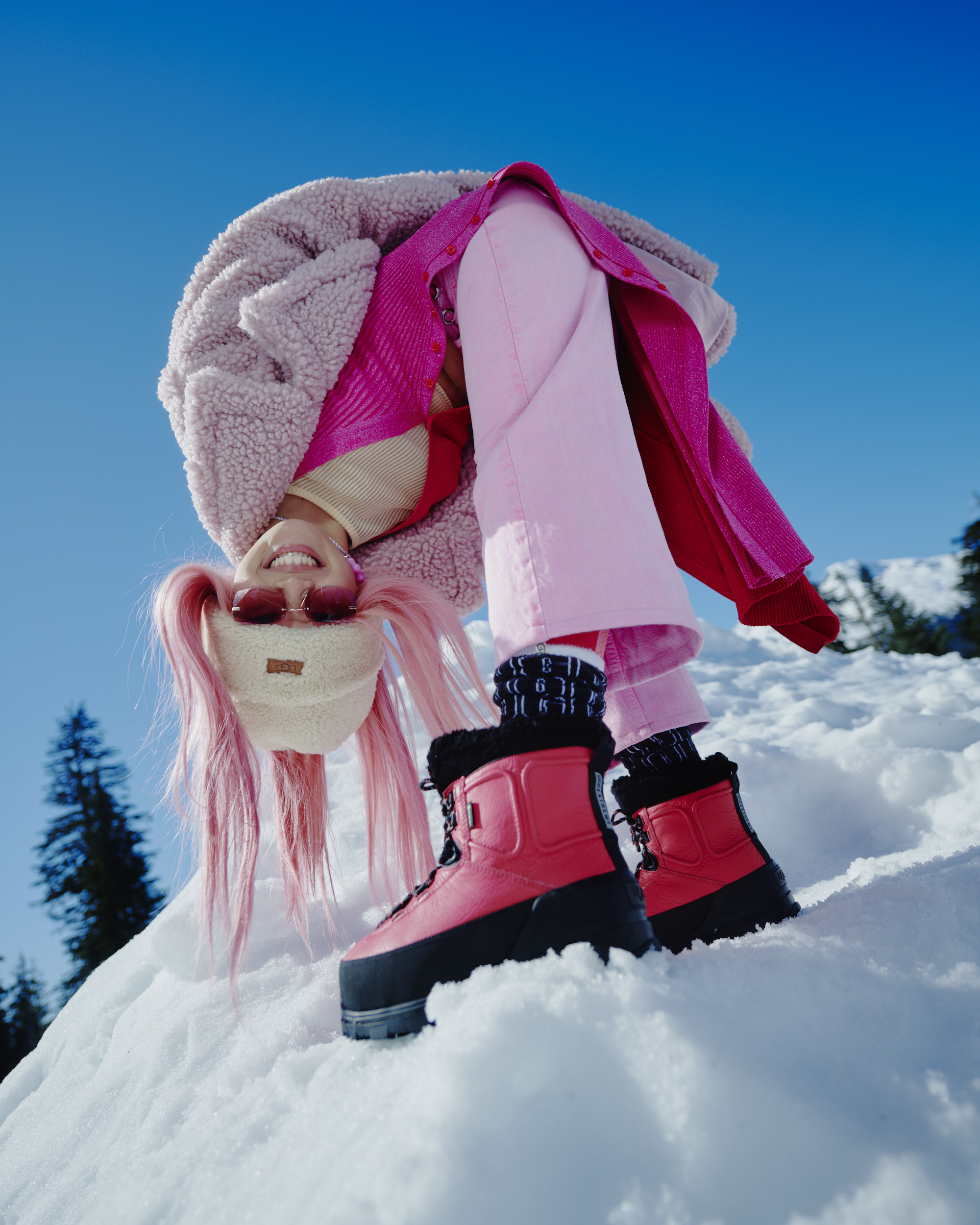 Woman in pink outerwear, hat, and red snow boots poses playfully on snow, set against a clear blue sky with scattered trees.