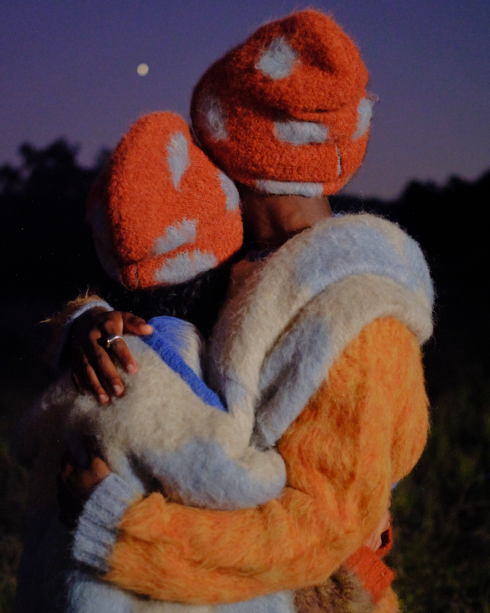 A couple wearing matching mushroom hats and fuzzy coats embraces in a field under the moonlit sky, creating a whimsical and cozy scene.