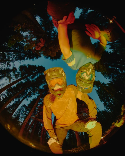 Two mountain bikers wearing helmets in a forest at dusk; fisheye lens captures dynamic, inverted perspective of the riders and tall trees.