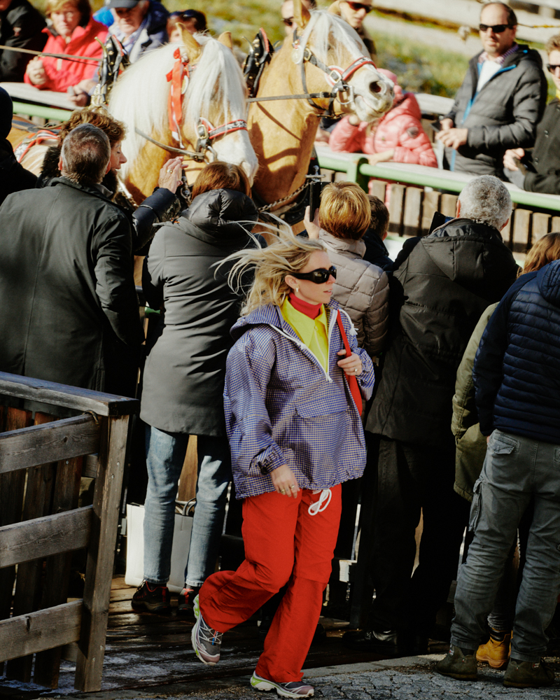 Blond woman in red pants walks past a group of people, with two horses and a carriage visible in the background during daytime.