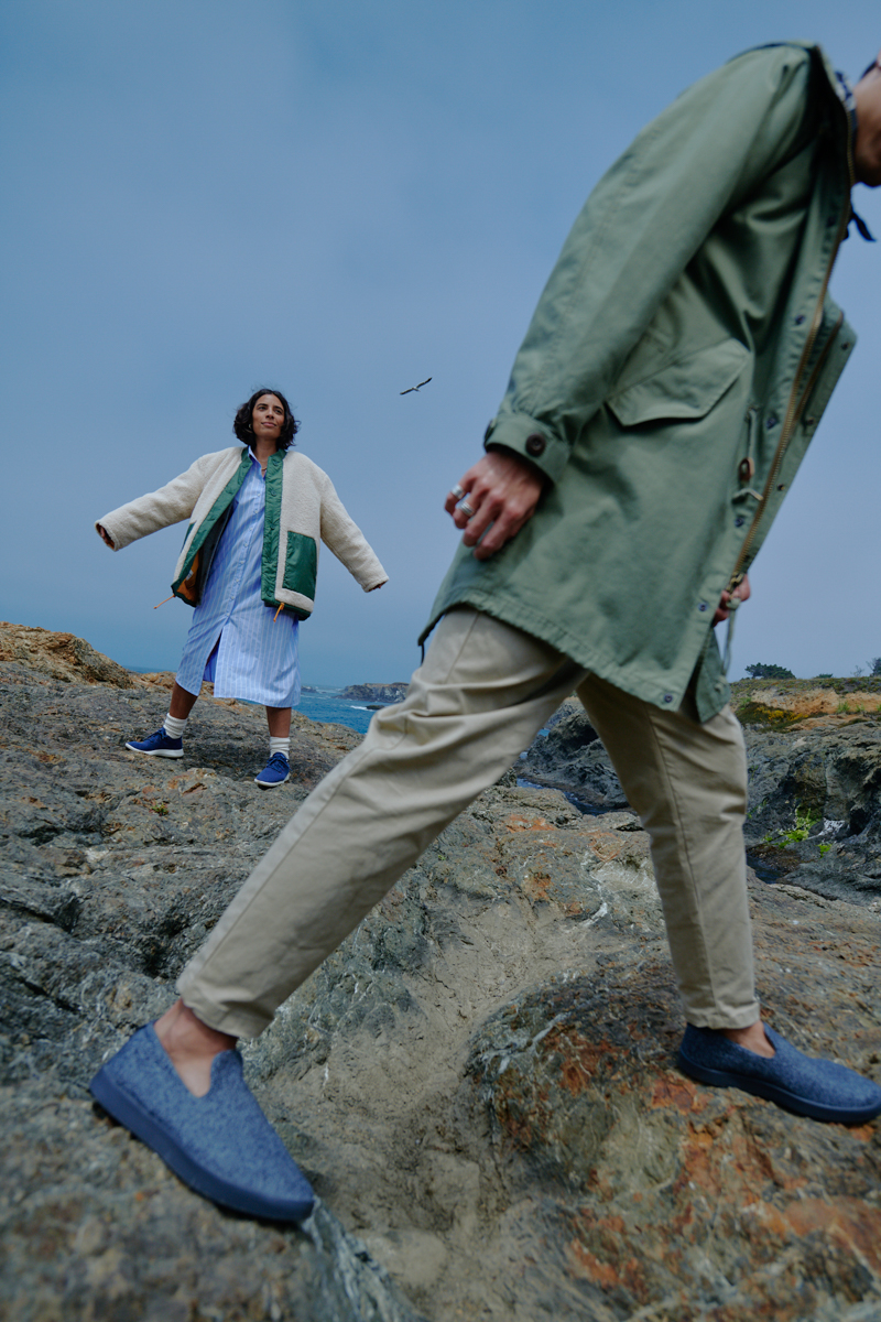 Two people posing on a rocky beach with cloudy sky. Woman wears jacket, man in coat and pants. Both wear slip-on shoes.