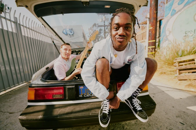 Two men posing together with a guitar inside the open hatch of a parked car on an asphalt street in Los Angeles.
