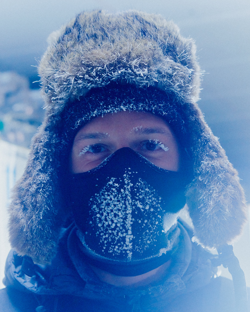 A close-up of a man in cold weather gear and a fur hat, with frost on his eyelashes and face mask, creating a wintery portrait.
