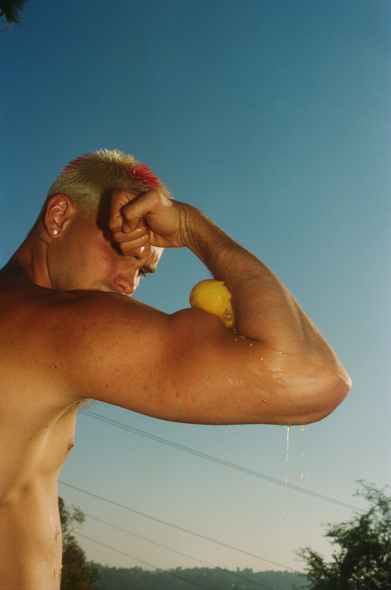 Adult man with a blonde mohawk presses a lemon against his arm, outside in the sunshine, set against a clear blue sky and greenery.