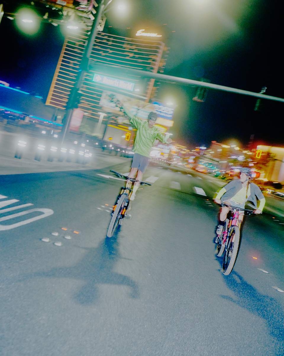 Two cyclists ride down a busy street at night in front of brightly lit buildings and traffic, with motion blur conveying movement.