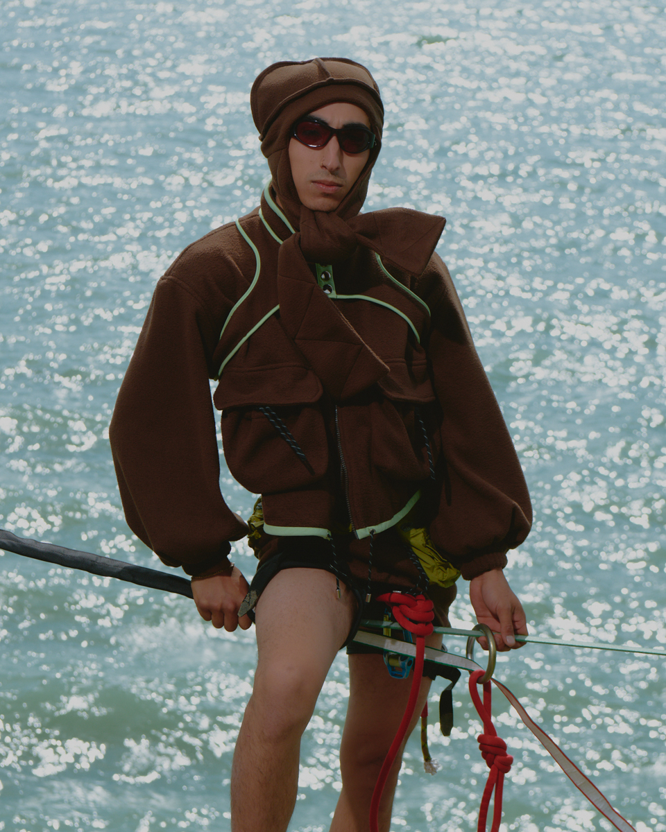 An adult male balances on a slackline, wearing a brown outfit and sunglasses, with a glimmering blue body of water in the background.