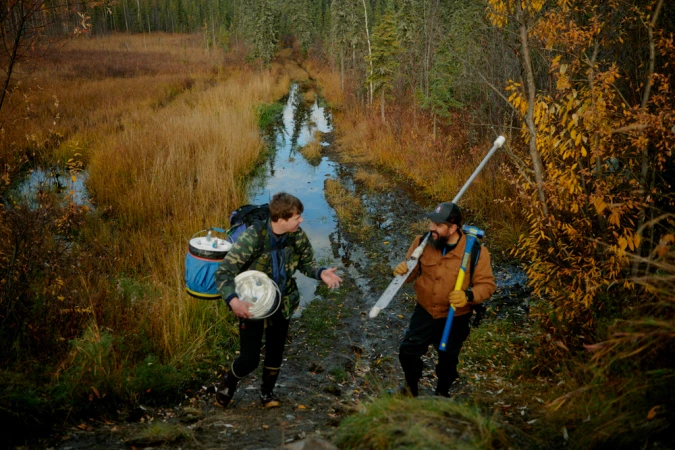 Two men carrying field equipment walk along a muddy path through a sparse northern forest landscape.