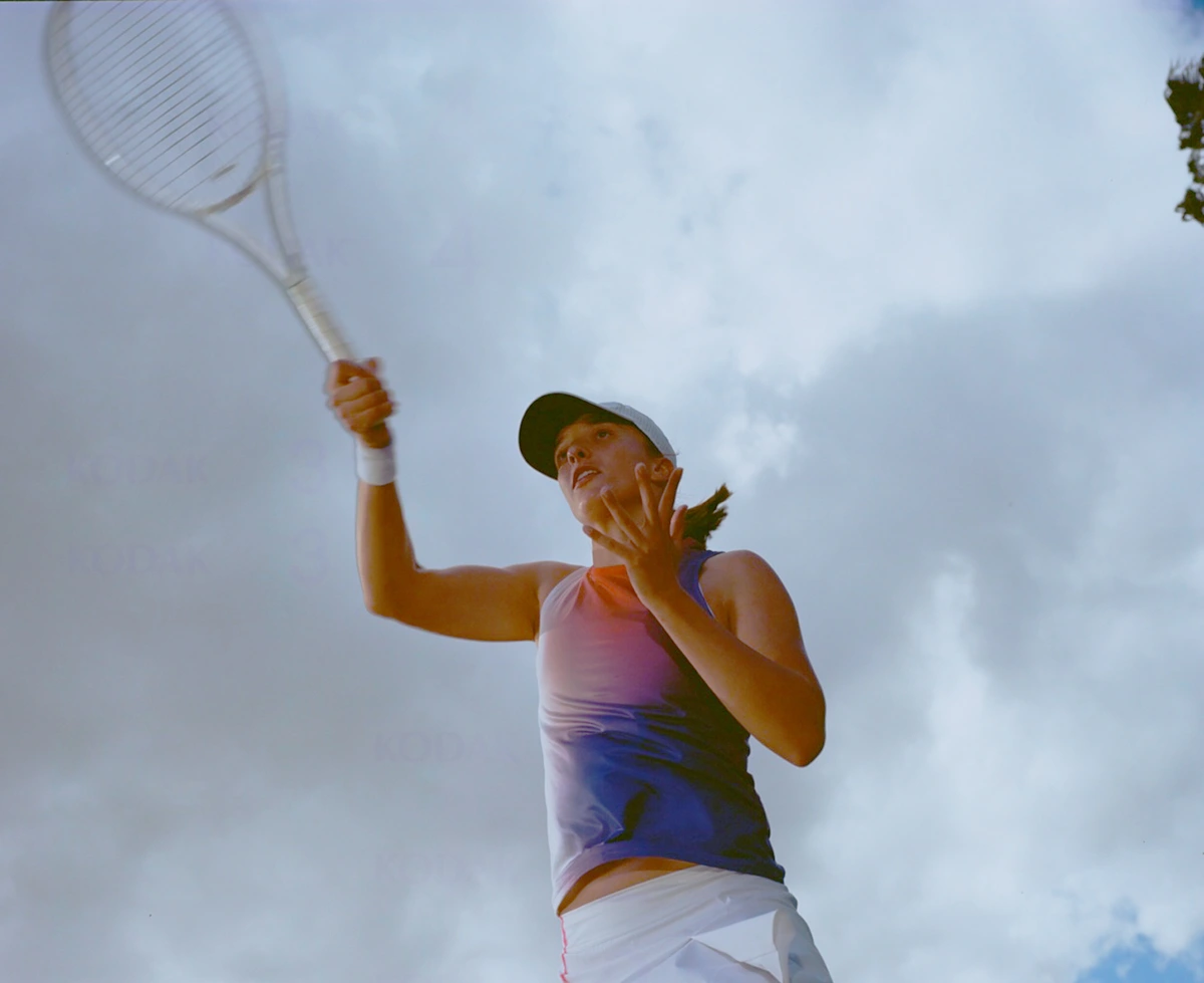 Female tennis player swinging a racket against an overcast sky, captured from a low angle by photographer Frankie Carino.