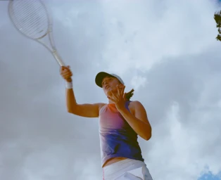 Female tennis player swinging a racket against an overcast sky, captured from a low angle by photographer Frankie Carino.