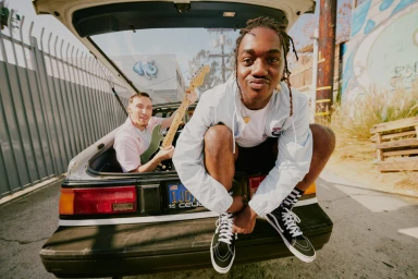 Two men posing together with a guitar inside the open hatch of a parked car on an asphalt street in Los Angeles.