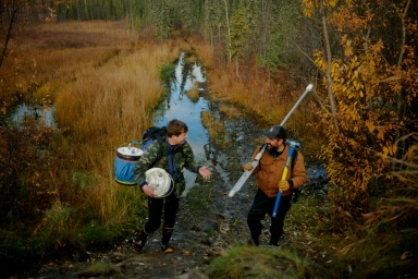Two men carrying field equipment walk along a muddy path through a sparse northern forest landscape.