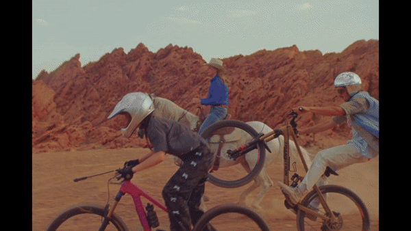 Three people riding mountain bikes and a horse across a dusty desert landscape with rugged red rock formations.