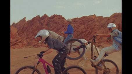 Three people riding mountain bikes and a horse across a dusty desert landscape with rugged red rock formations.