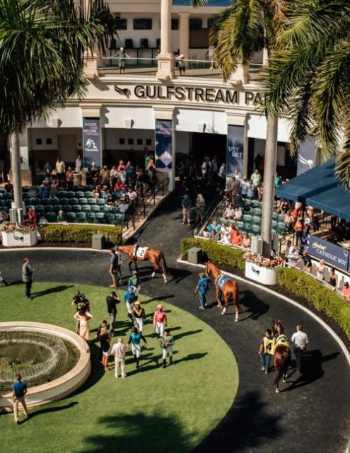 Outdoor daytime view of jockeys, horses, and spectators at Gulfstream Park horse racing track with palm trees and architecture.
