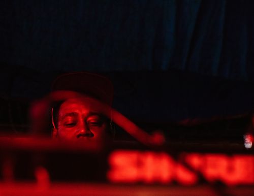 Adult male with dark skin and closed eyes wearing a cap in a dark, indoor environment with red lighting and musical equipment.