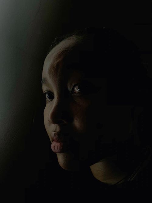 Portrait of a young Black woman with a neutral expression, looking off to the side, set against a dark, shadowed background.