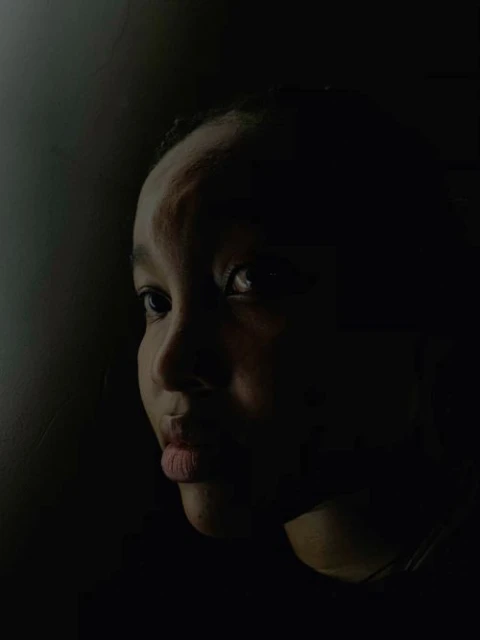 Portrait of a young Black woman with a neutral expression, looking off to the side, set against a dark, shadowed background.