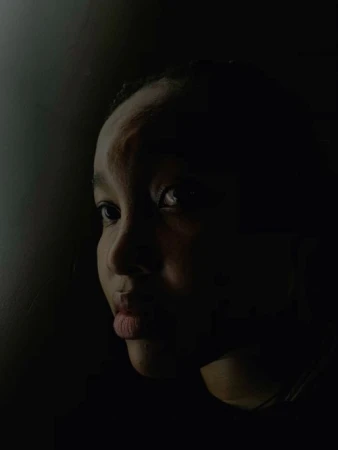 Portrait of a young Black woman with a neutral expression, looking off to the side, set against a dark, shadowed background.