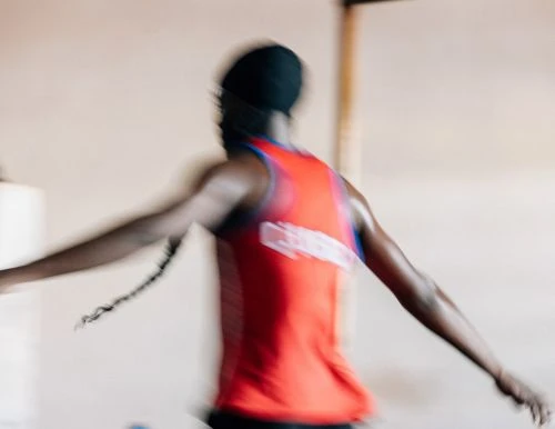 Motion blur of an adult Black man wearing a red sleeveless shirt and a black cap indoors.