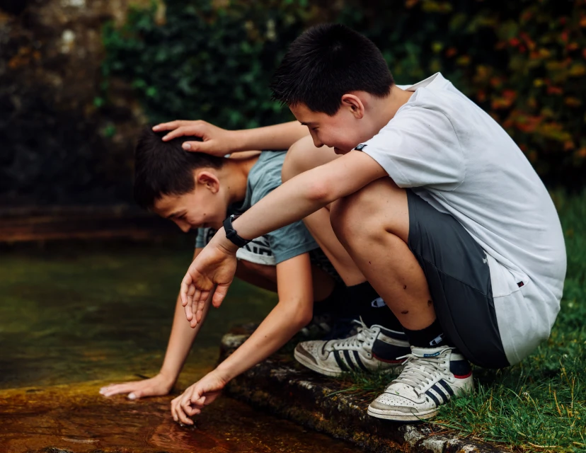 Two boys squat outdoors by a body of water; one places a hand on the other's head in a natural setting with greenery and foliage.
