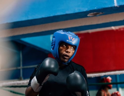 Female boxer in blue headgear and black gloves inside a boxing ring looking up, ready for a match in an indoor environment.