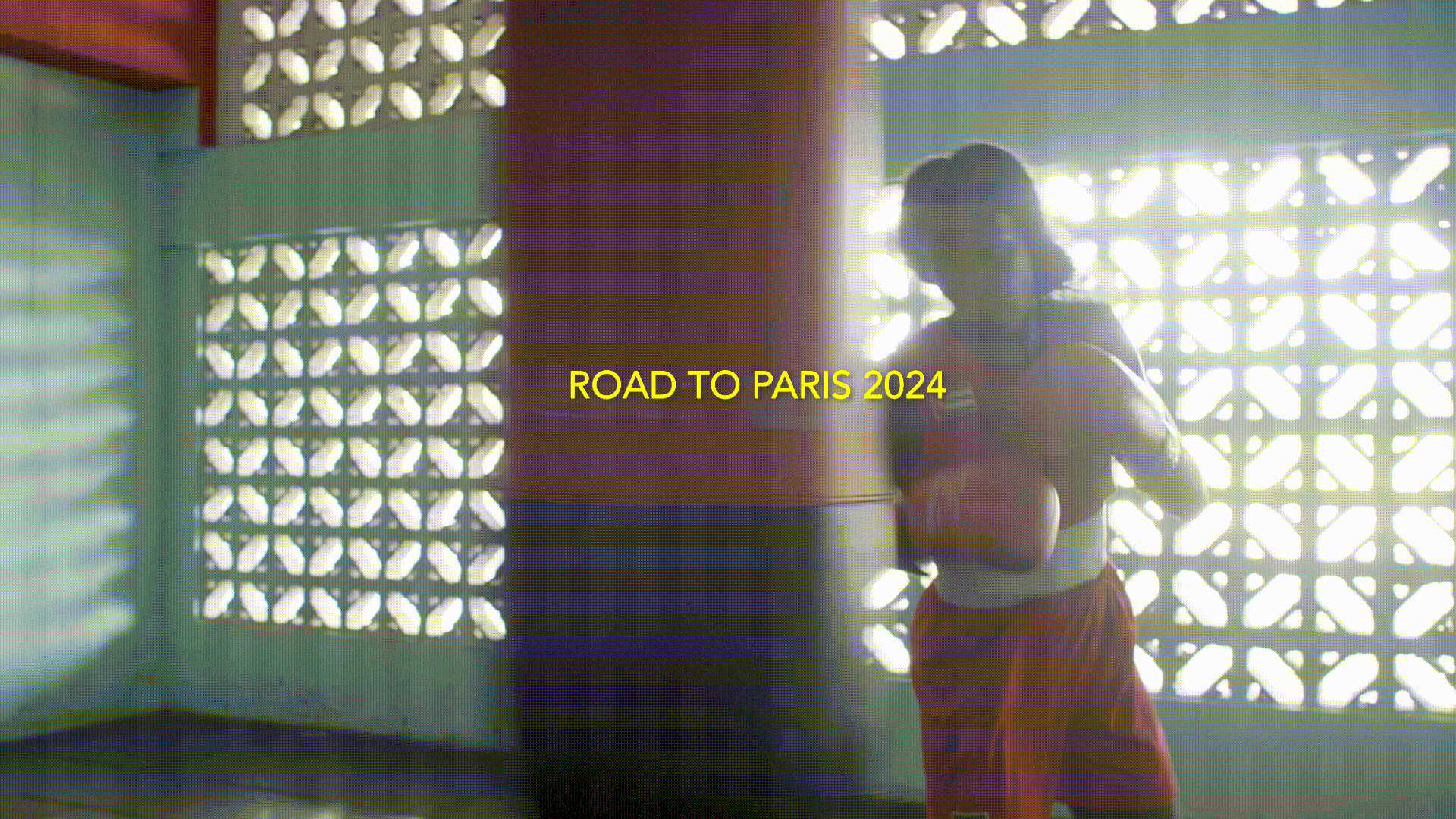 A boxer wearing red shorts and boxing gloves stands next to a punching bag in a brightly lit gym with vintage-style windows.