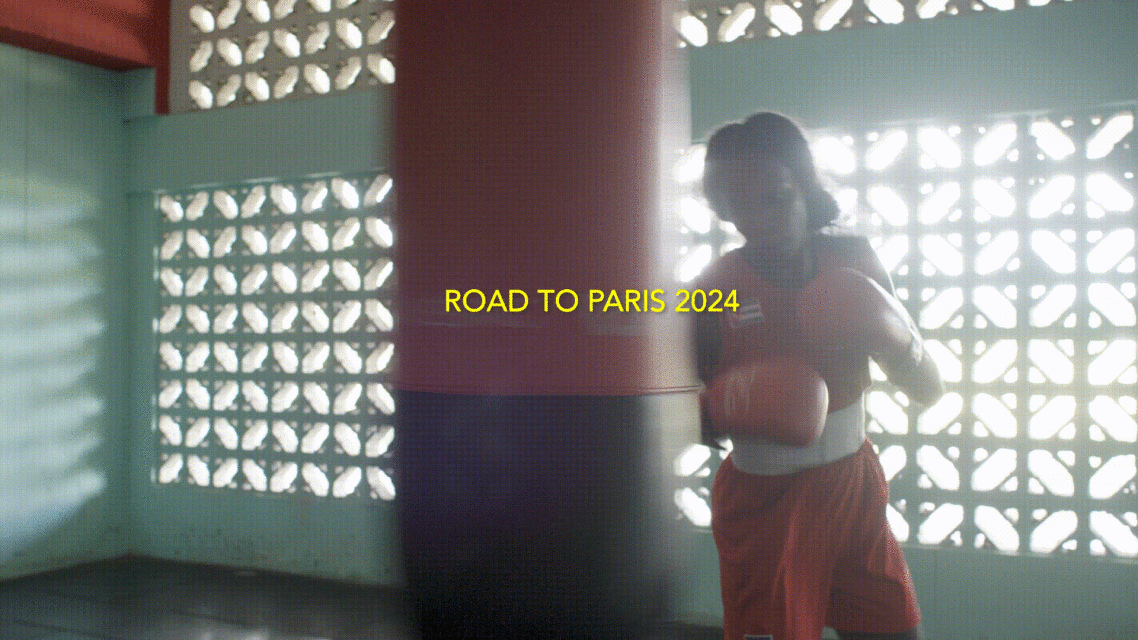 A boxer wearing red shorts and boxing gloves stands next to a punching bag in a brightly lit gym with vintage-style windows.