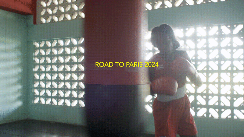 A boxer wearing red shorts and boxing gloves stands next to a punching bag in a brightly lit gym with vintage-style windows.
