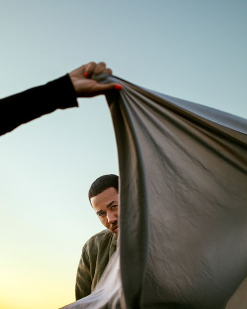 A man peeks from behind a gray fabric, held up by a hand with red nail polish against a serene blue sky at golden hour.