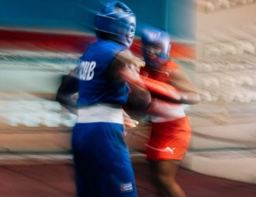 Two people compete in a boxing match, both wearing protective headgear and boxing gloves, set against a blurred indoor background.