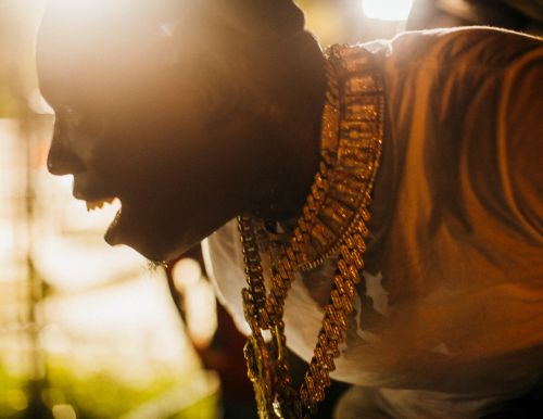 Profile shot of an adult man wearing gold chains and a light-colored shirt, illuminated by bright sunlight, showing an open mouth.