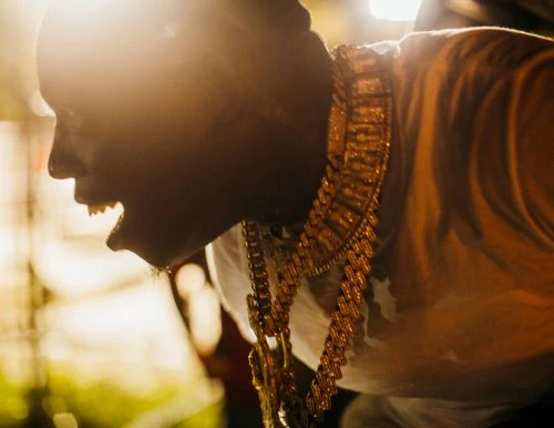 Profile shot of an adult man wearing gold chains and a light-colored shirt, illuminated by bright sunlight, showing an open mouth.