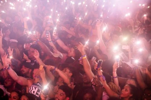Large crowd of people holding up phones with flashlights in a dark venue at a concert or event, creating a vibrant and energetic atmosphere.