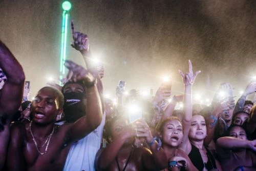 Enthusiastic crowd with cell phones at concert, showcasing diverse group of people cheering, illuminated by stage lighting in an outdoor setting.