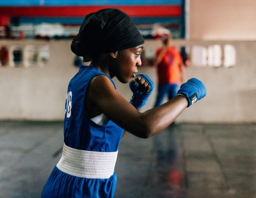 Female boxer in blue uniform and boxing gloves throwing a punch in an indoor boxing gym with another person in the background.