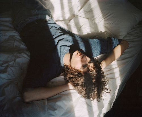 Adult lying on a white bed in a room with light and shadow, looking upwards with arms above their head, hair spread across the pillow.
