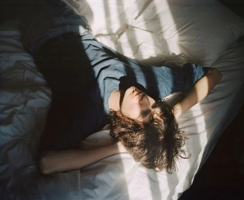 Adult lying on a white bed in a room with light and shadow, looking upwards with arms above their head, hair spread across the pillow.