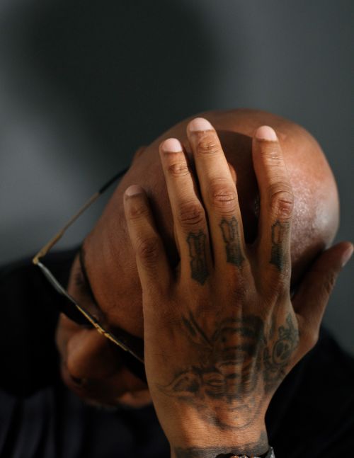 Close-up studio shot of a bald adult with a tattooed hand covering part of their face, also wearing glasses, set against a grey backdrop.