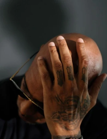 Close-up studio shot of a bald adult with a tattooed hand covering part of their face, also wearing glasses, set against a grey backdrop.