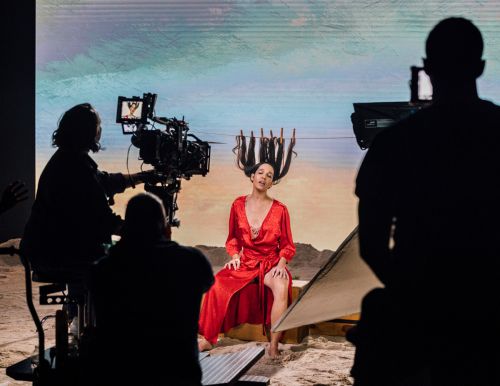 Adult female model in a red dress with a desert landscape background, photographed with film crew in a photo studio.
