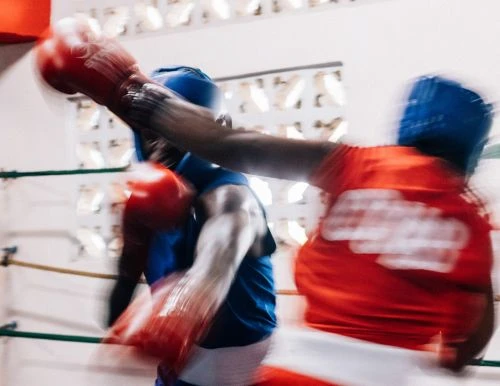 Two boxers, one in blue, one in red, compete in a boxing ring, showcasing athleticism in a dynamic sports scene with blurred motion.