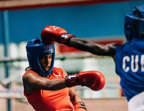 Two boxers in a boxing ring, one in a red top and blue headgear throwing a punch at another boxer in blue uniform.
