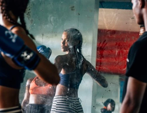 Woman trains in a boxing gym, reflecting in the mirror. She wears gloves and boxing attire, in an indoor setting.