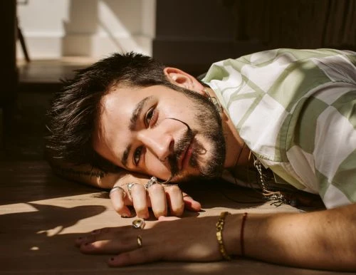 Close-up of a Hispanic adult male lying on a wooden floor, wearing a patterned shirt, bracelets, and rings, looking at the camera.